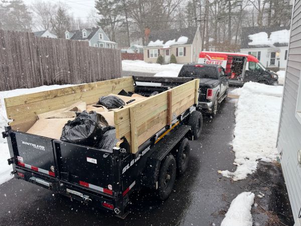 Snow falling on a trailer filled with bags and rolled carpet in a residential driveway.