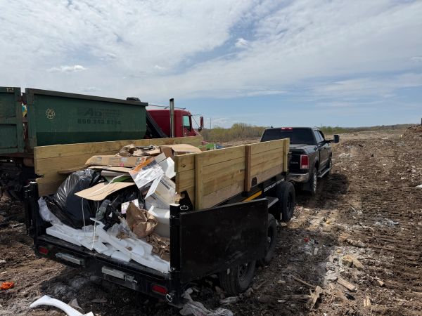 A black pickup truck with a wooden trailer filled with trash at a landfill site.