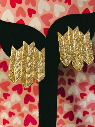 Gold and rhinestone arrow-shaped earrings on a black stand with heart-patterned background.