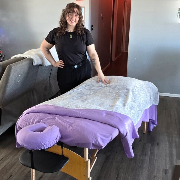Smiling woman stands beside a massage table with lavender sheets in a cozy room. Mobile massage
