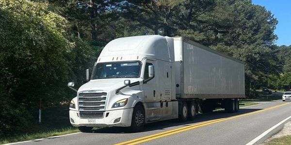 White semi-truck driving on a rural road surrounded by trees.