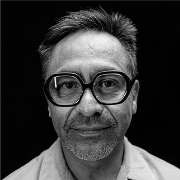 Black and white portrait of a man with large glasses against a dark background.