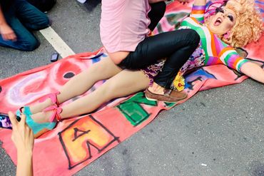A colorful performer lies on a rainbow banner during a pride event, being photographed.