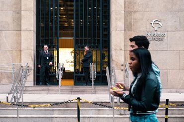 Two security guards stand at the entrance of a bank with two pedestrians walking past.
