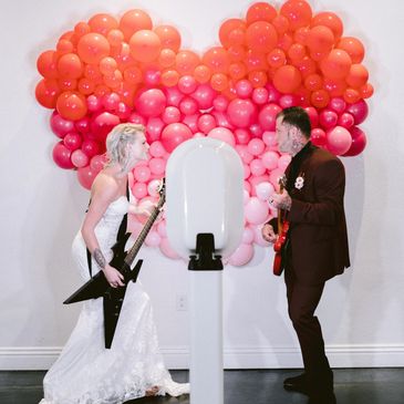 Bride and groom play guitars in front of heart-shaped balloons.