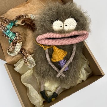 A fuzzy crochet creature with big eyes and long lips in a box.
