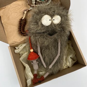 A fuzzy, handmade creature with big eyes and crocheted limbs in a cardboard box.