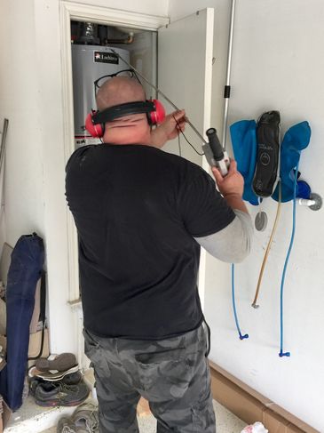 A plumber has on earphones and is holding a long tool up to a water heater, doing leak detection