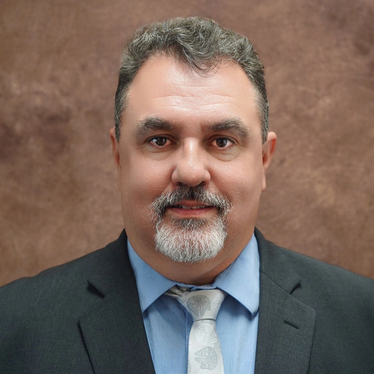 A middle-aged man in a suit and tie smiling gently at the camera.