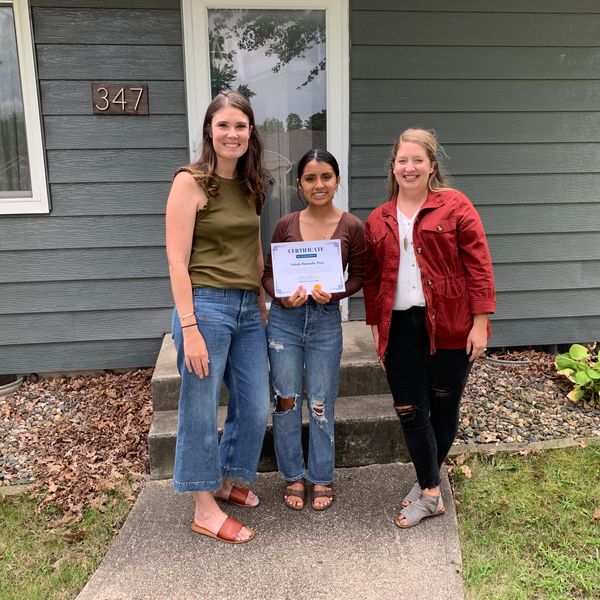 Three women standing on a porch, one holding a certificate.