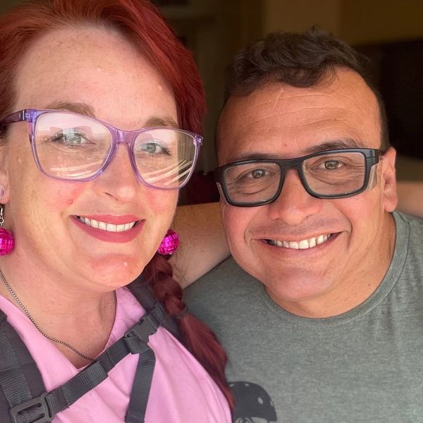 Two smiling people wearing glasses, one with red hair and pink earrings.