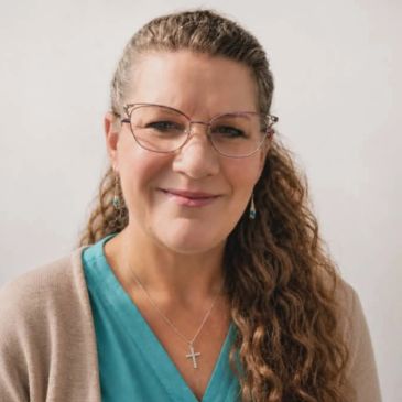 Smiling woman with glasses and curly hair wearing a teal top and beige cardigan.