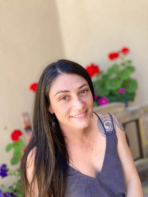 Smiling woman with long dark hair and a tattoo on her shoulder outdoors with flowers.