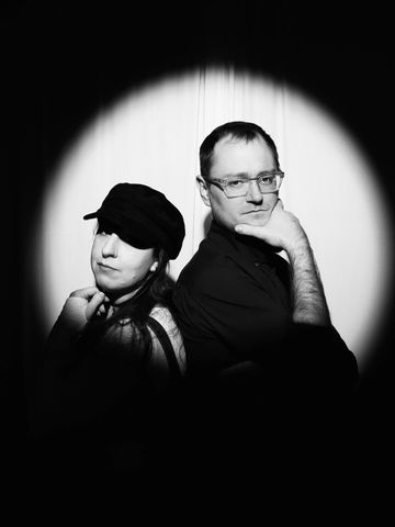 Two people pose back-to-back under a spotlight in black and white.