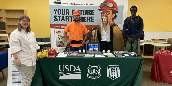 Four people stand behind a USDA Forest Service Job Corps recruiting table.