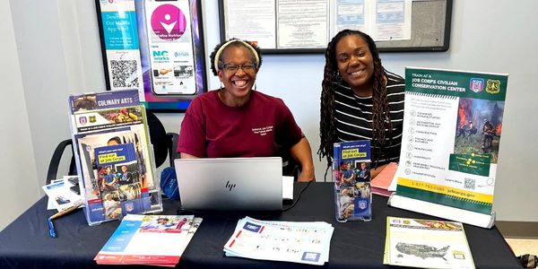 Two smiling women at a job training information table with brochures and laptop.