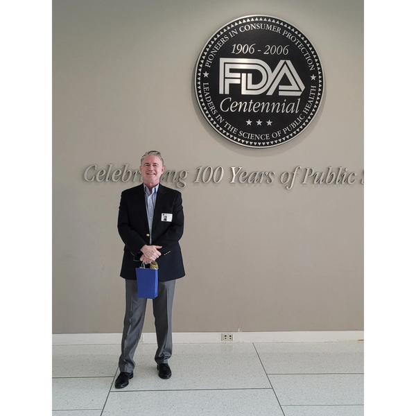 Man in formal attire standing in front of FDA Centennial wall plaque.