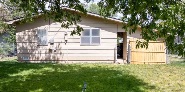 Backyard view of a beige house with green lawn and leafy tree branches above.