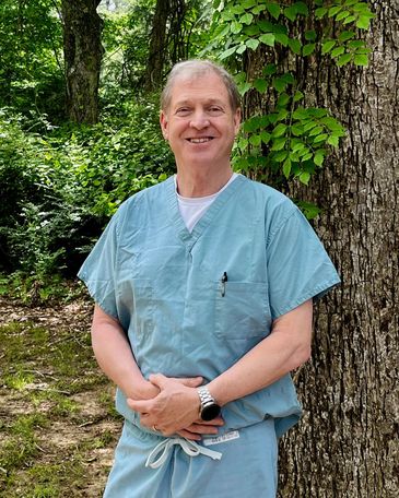 Dr. Phillip Green wearing medical scrubs and standing in front of a tree smiling.