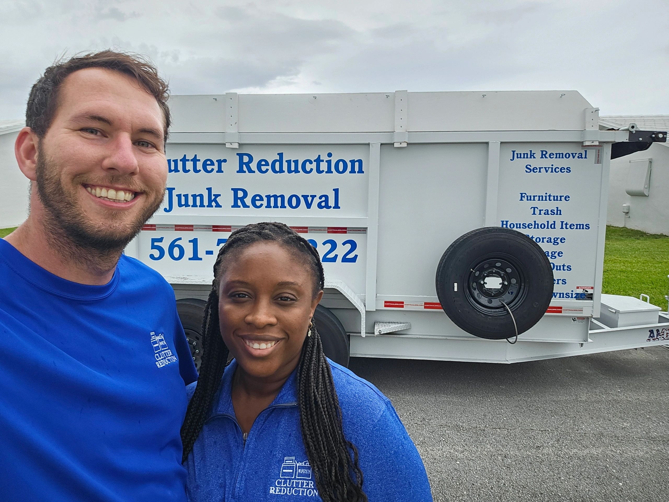 Two smiling people in blue Clutter Reduction shirts with a junk removal trailer behind them.