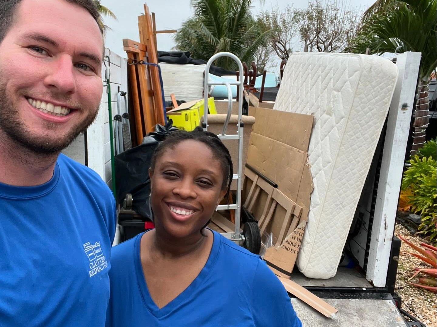 Two smiling people in blue shirts with a truck full of furniture and boxes behind them.