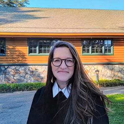 Smiling woman with glasses stands outdoors in front of a rustic house.