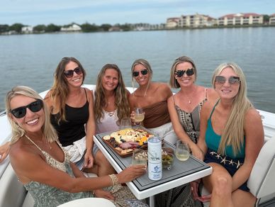 Six women enjoying drinks and snacks on a boat by the water.