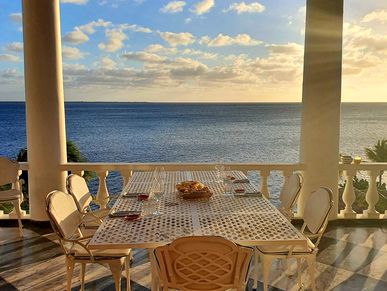 Outdoor dining table set with chairs overlooking the ocean at sunset.