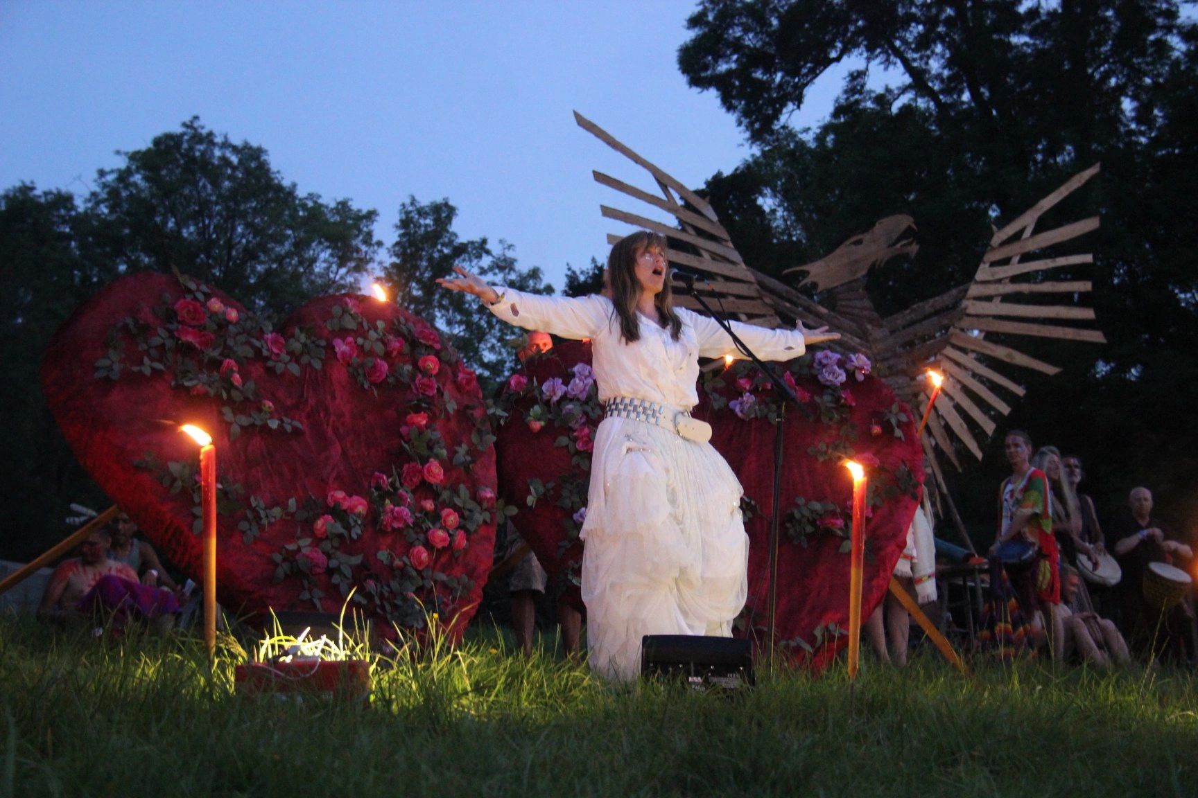 A woman in white performs outdoors with heart and eagle decorations at dusk.