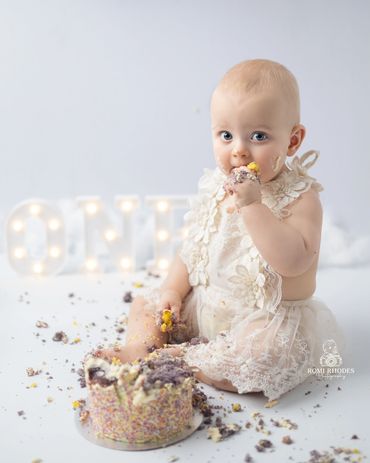 A baby in a white dress enjoying a messy first birthday cake.