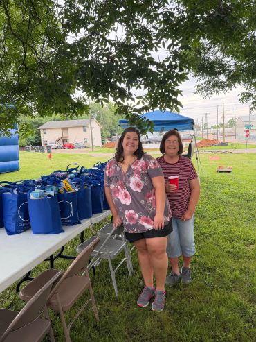 Two women smiling in a park near a table with blue bags.