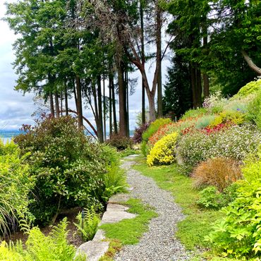 A winding gravel path through a vibrant garden with colorful flowers and tall trees.