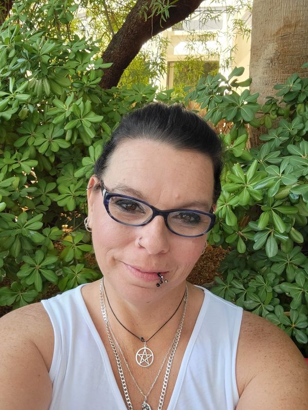 Woman with glasses and lip piercing smiles outdoors in front of green foliage.