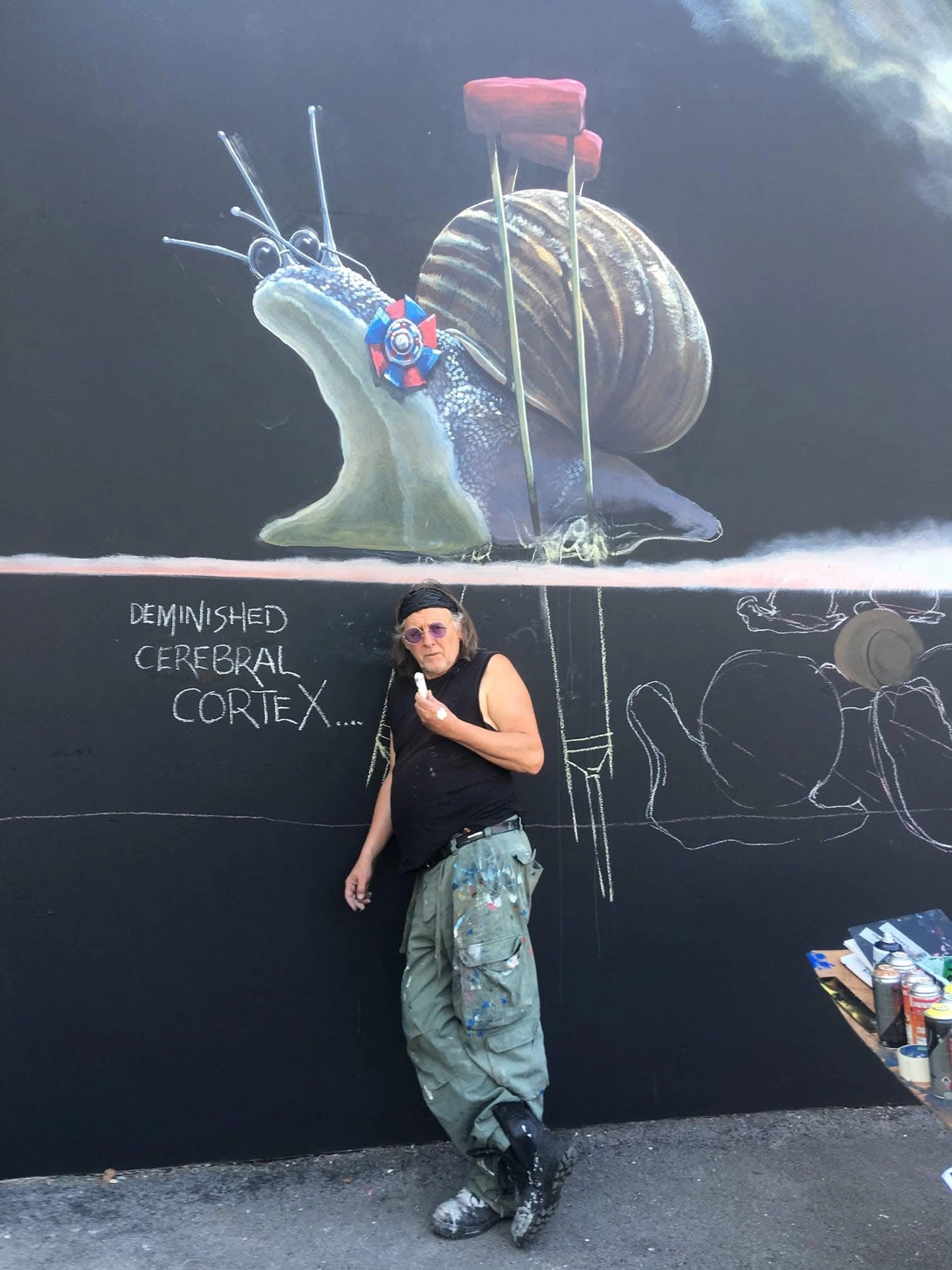 Artist posing in front of a mural of a snail with crutches and brain injury text.