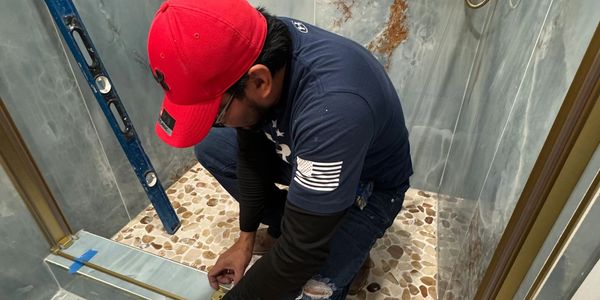 A man wearing a red cap installs a glass shower door in a tiled bathroom.