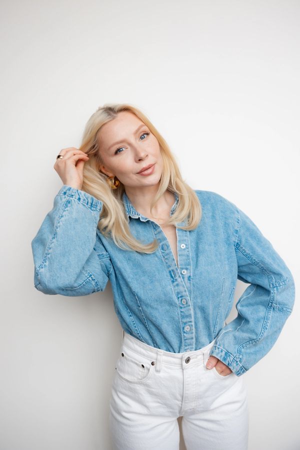 Blonde woman in denim shirt and white pants poses against white background.