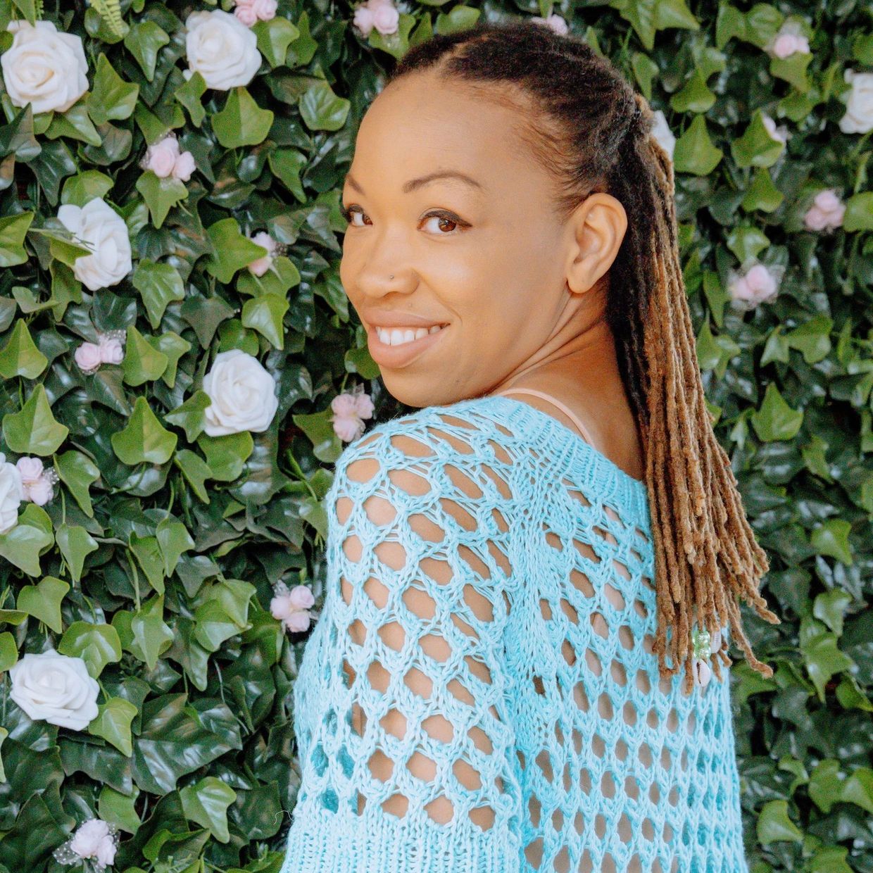 Smiling woman with dreadlocks wearing a blue crochet top in front of a floral wall.