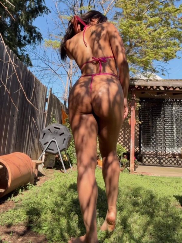 Woman in pink bikini walking barefoot in a sunlit garden with shadows of branches.