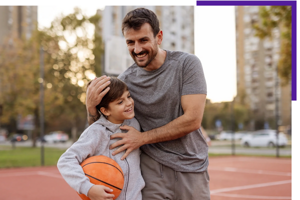 Father and son smiling on a basketball court with a ball.