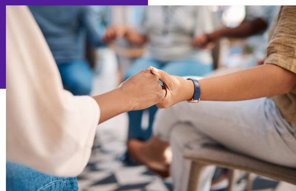 Two people holding hands in a supportive gesture during a group meeting.