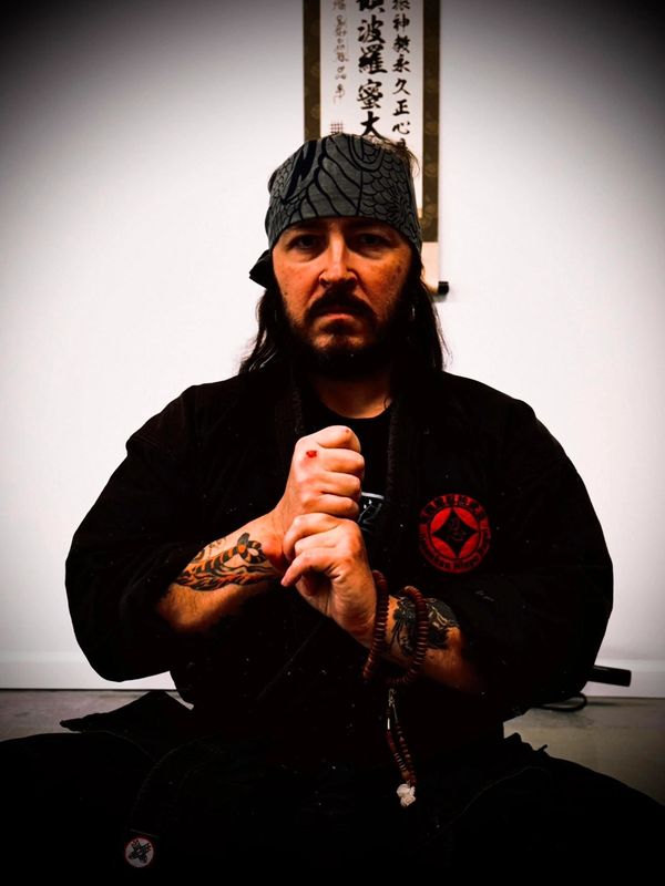 Martial artist in black gi with tattoos and bandana poses seriously indoors.