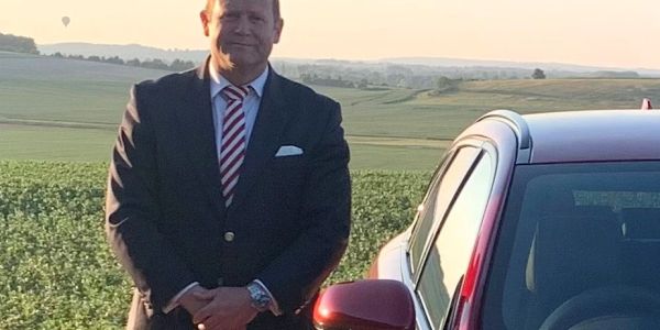 Man in suit stands next to a red car in a scenic rural area at sunset.
