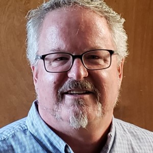Smiling man with glasses and gray hair wearing a blue checkered shirt.