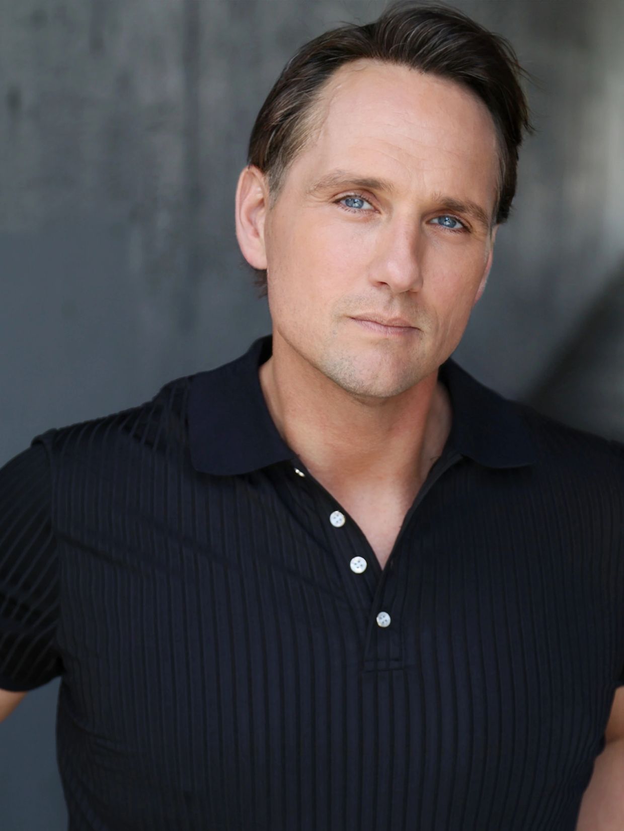Man with blue eyes wearing a black polo shirt, posing against a gray background.