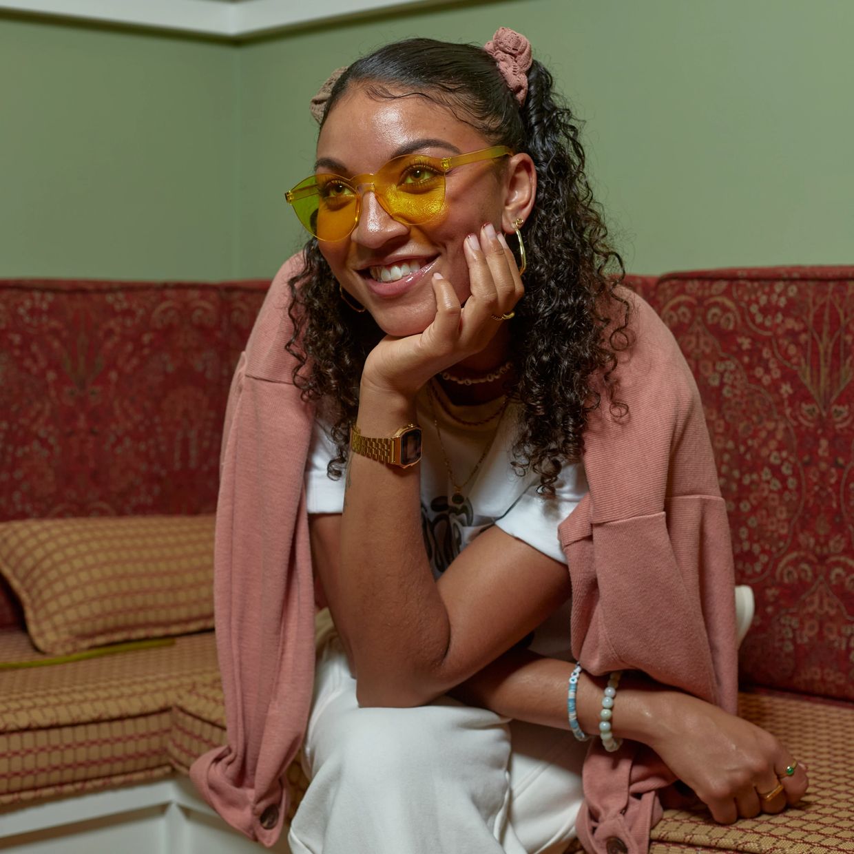 A smiling woman with curly hair and yellow glasses sitting on a red patterned sofa.