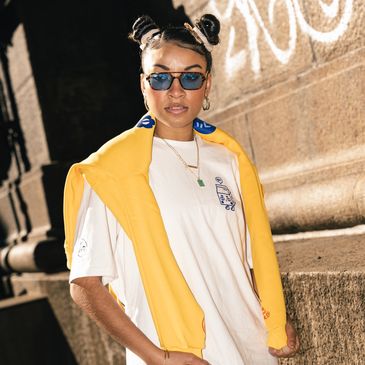 Trendy woman with sunglasses and space buns poses with a yellow jacket over her shoulders.