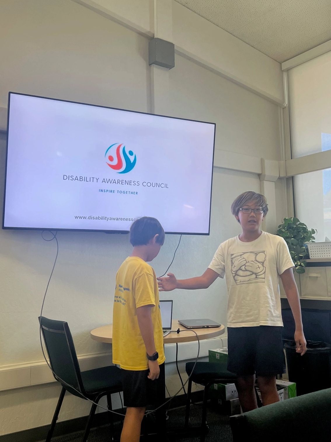 Two boys standing in a room with a Disability Awareness Council presentation on screen.