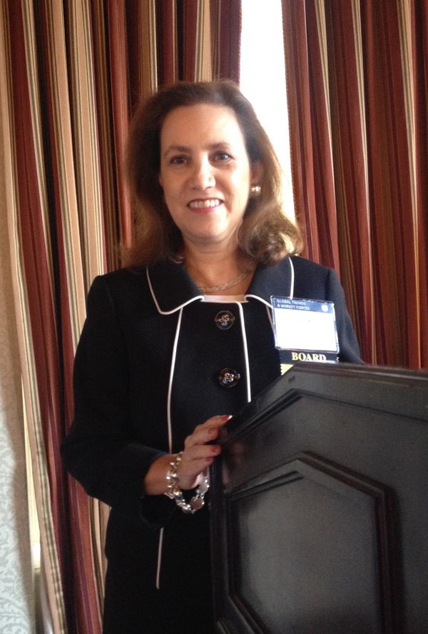 Smiling woman in formal black suit standing behind a podium indoors.