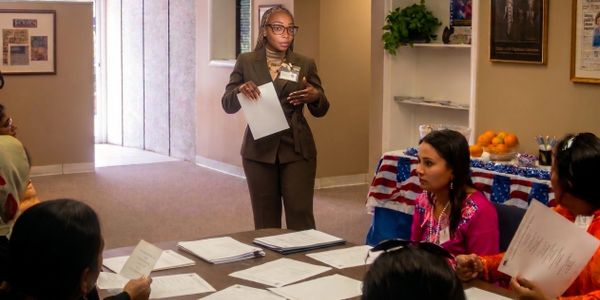 A woman stands presenting to a seated group with papers in a meeting room.