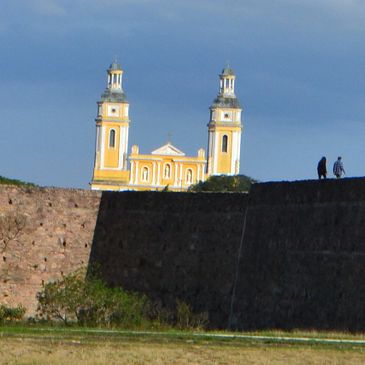 Historic fortress wall with two people walking and a yellow church in the background.